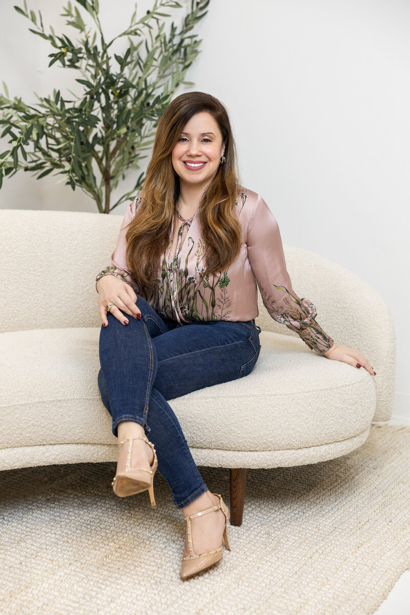 Kathy Gladys Colon sitting on a cream-colored couch with a welcoming smile, surrounded by greenery