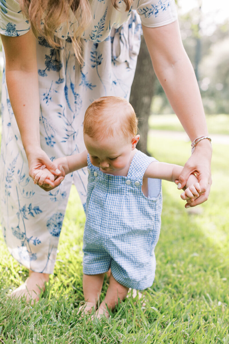 Mom leaning down holding baby's arms while he stands in the grass
