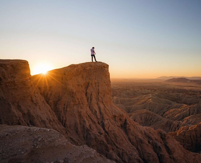 Traveler standing atop a breathtaking cliff at sunset, overlooking a vast desert landscape—part of a luxury overland adventure to hidden locations