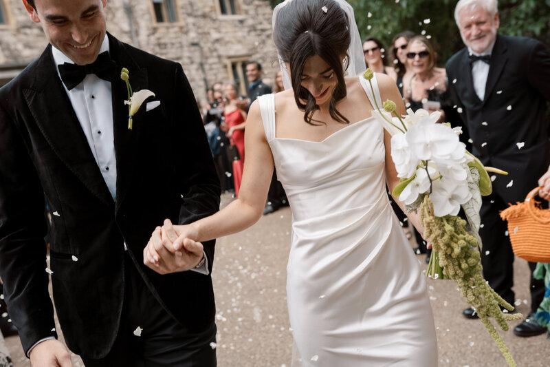 Bride and groom walk hand in hand surrounded by wedding guests celebrating with confetti.