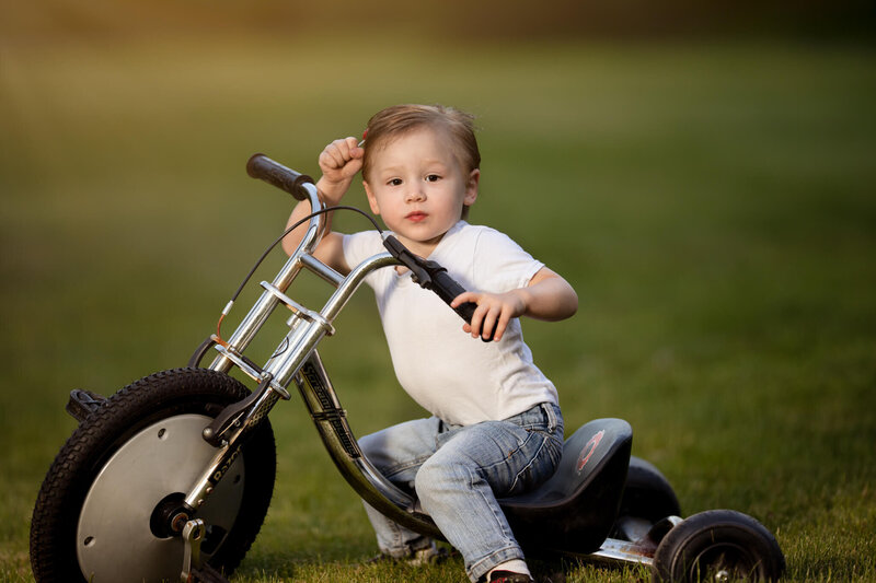 Toddler boy holding a lollipop in his heair while sitting on a tricycle greaser style hair and outfit