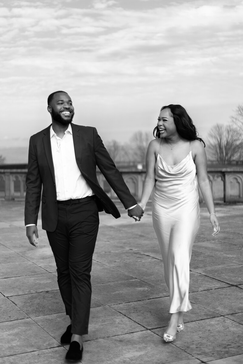 Ochre Court Mansion  | Kelsey Sheehan Photography Timeless Rhode Island Weddings | A joyful couple walks hand in hand on a paved path. The man wears a suit, and the woman wears a flowing dress. They are both smiling and laughing.