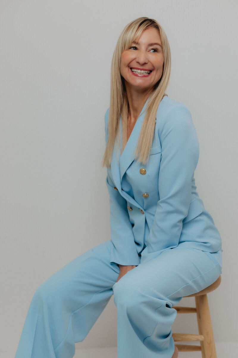 A women in a light blue power suite sitting on a stool smiling. 