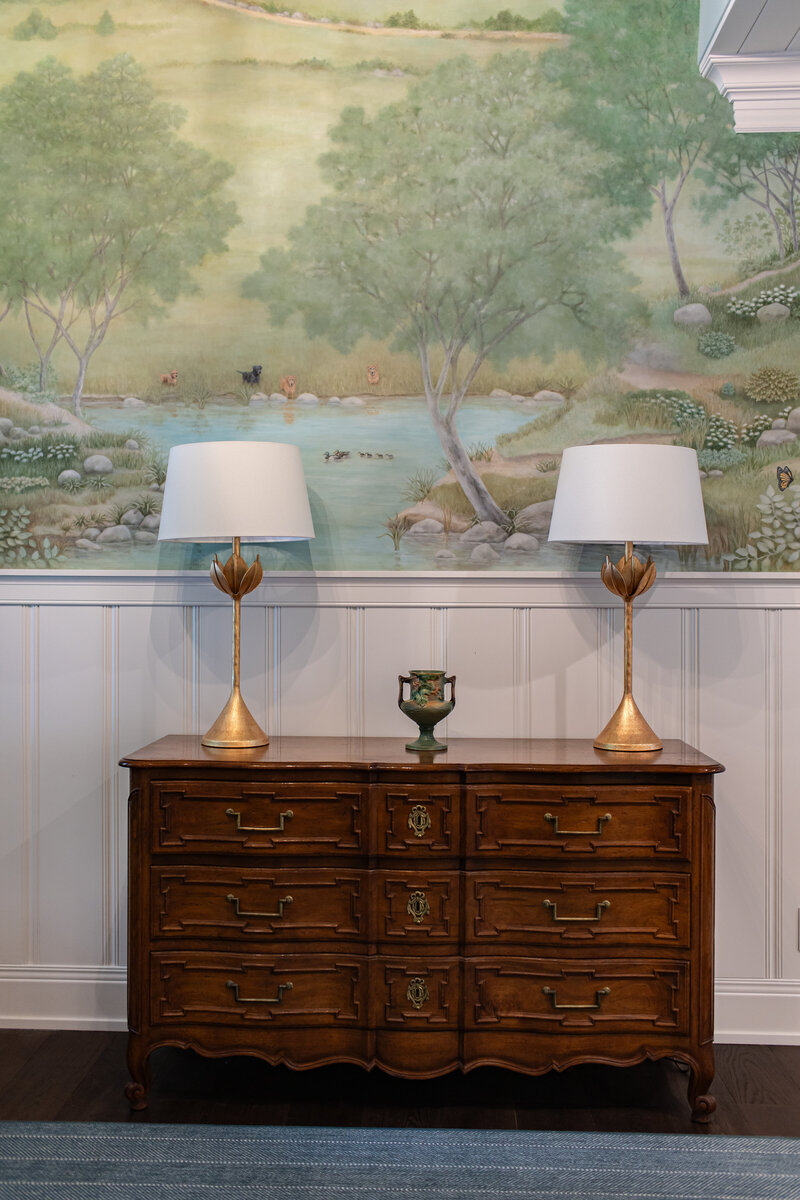 A stylish entryway featuring custom wallpaper, detailed wall paneling, and a wooden entry table accented by gold lamps. The space exudes sophistication and warmth, setting the tone for a beautifully designed home.