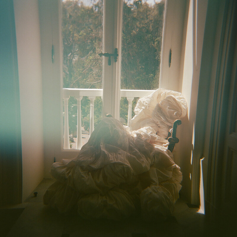 Voluminous wedding gown cascading over a vintage chair beside a sunlit balcony window, captured with ethereal light and film haze