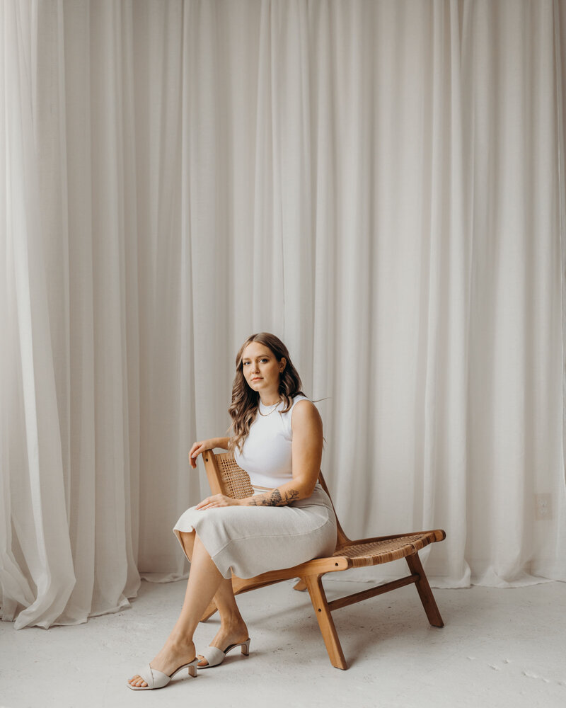 Brand and Web Design in Hamilton Ontario sitting on a wicker and wood chair in front of a linen curtain in a neutral photo studio