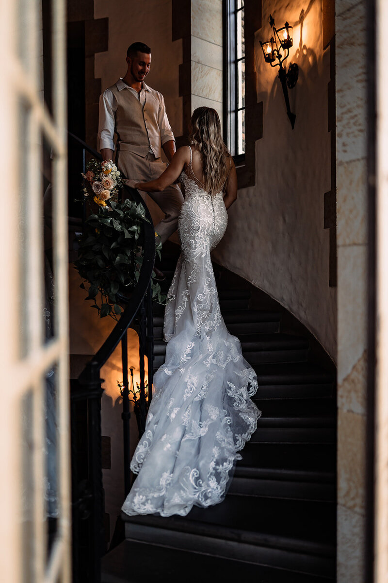 Bride walking up the stairs while her groom walks down to meet her on a winding staircase indoors with soft lanterns on the walls