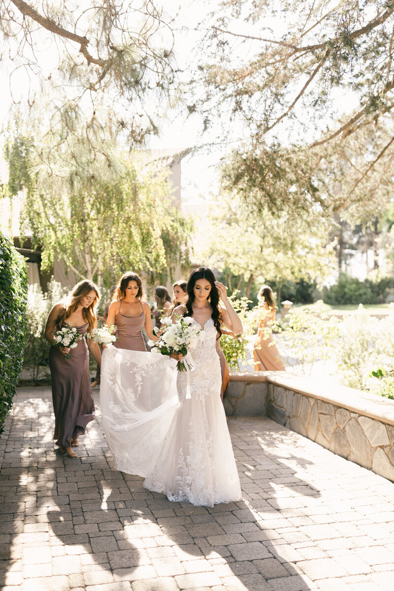 Elegant wedding moment at Maravilla Gardens in Los Angeles, CA — bride walks with bridesmaids in neutral-toned gowns under sunlit trees, captured in editorial style with fashion-forward details and natural movement.
