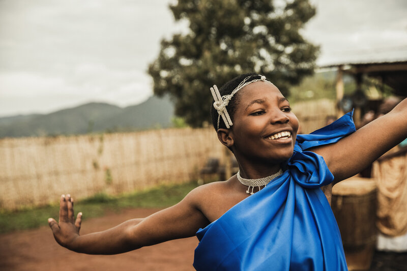 A young Rwandan woman in a vibrant blue traditional dress smiles joyfully while performing a cultural dance in a local village. Connect with the community by spending a day in a traditional Rwandan village, engaging in local activities and immersive experiences. 