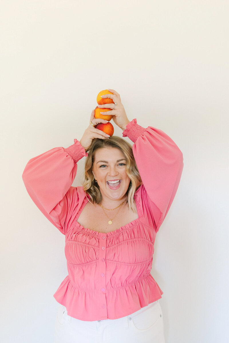 Mollie Mason in a pink top standing in front of a white wall holding a stack of 3 oranges on her head