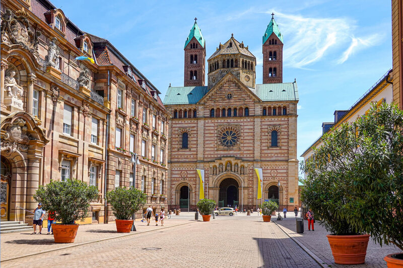 A large Romanesque-style cathedral with twin green-roofed towers facing a pedestrian street lined with historic buildings and potted trees.