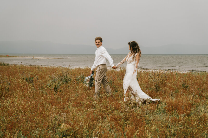 Groom holding bouquet in one hand and bride's hand in the other leading the bride on a walk through grass in front of a lake taken by California elopement photographer Kasey Mantiply