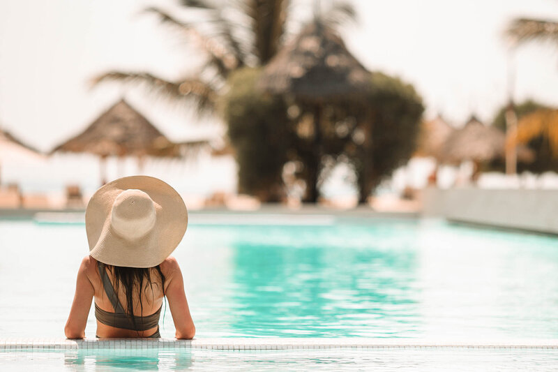 A woman wearing a wide-brimmed hat relaxes at the edge of a swimming pool, looking out toward palm trees and thatched umbrellas in the background on a sunny day.