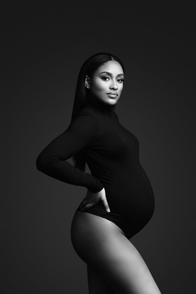 A striking black-and-white maternity portrait of a woman confidently posing in a form-fitting black outfit. The minimalistic background highlights her elegance and strength, capturing the beauty of pregnancy with a timeless and sophisticated touch.