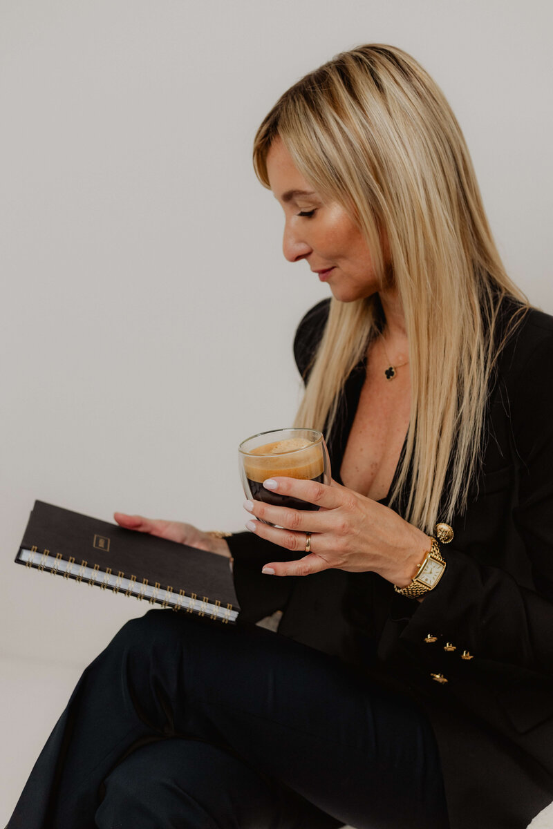 A women in black suite holding a coffee looking at a book.