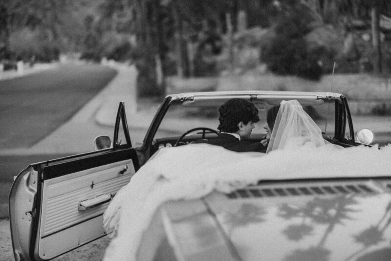 A timeless black and white wedding portrait of a newlywed couple in a vintage convertible, captured in Palm Springs, California. The bride's veil drapes over the car as they share an intimate moment, evoking old Hollywood romance with a modern love story.