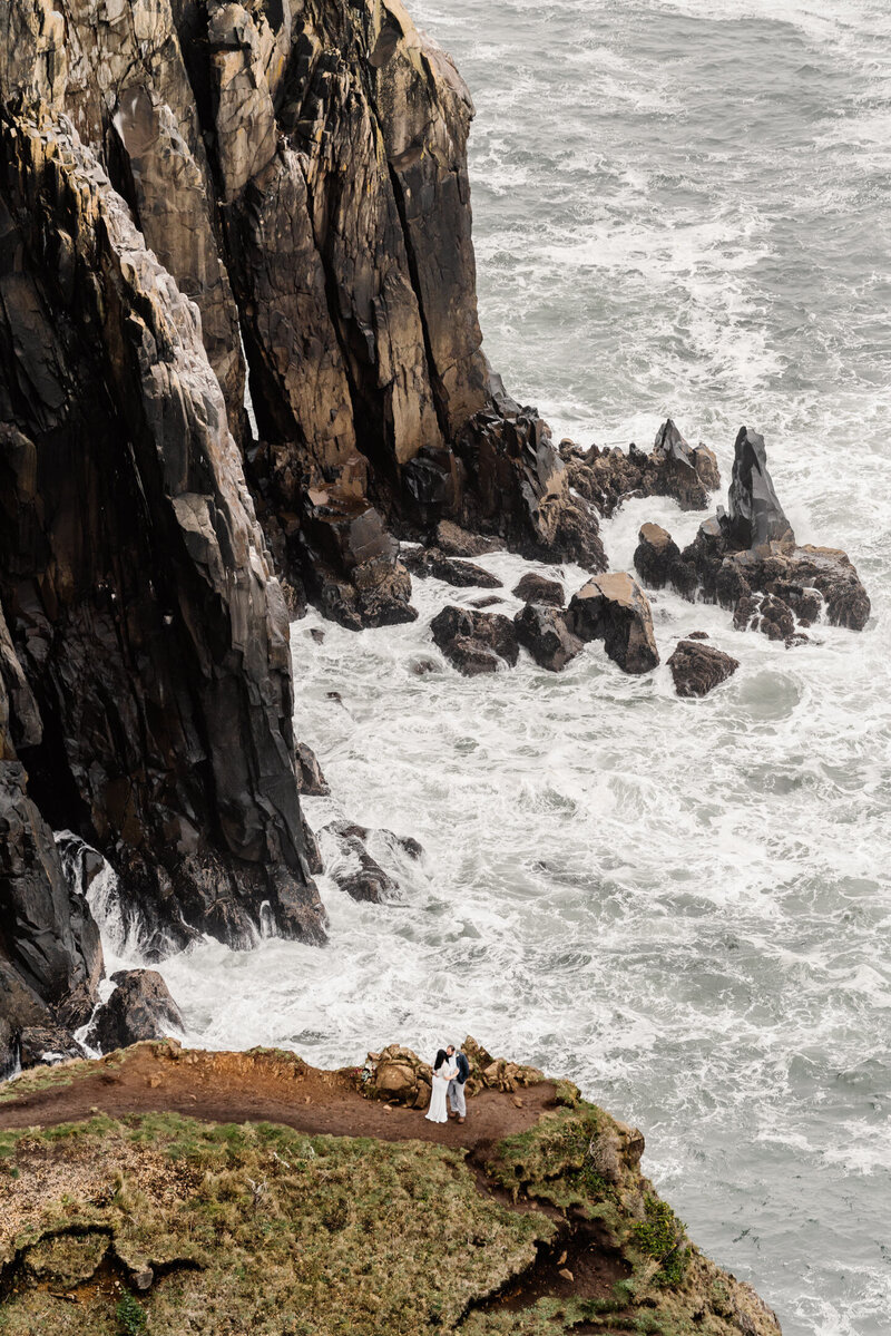 During their Oregon coast elopement, a bride and groom embrace at the edge of a coastal cliff, the blue sea churning white foam beneath them.