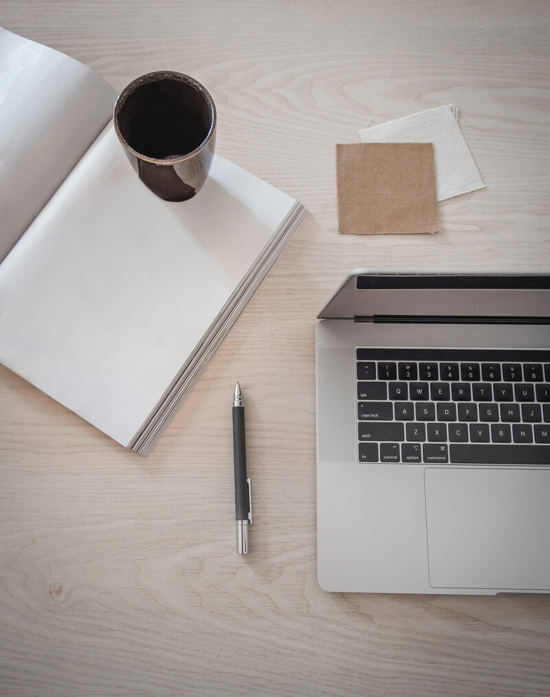 Overhead view of tan table top with open laptop, pen, notebook and brown coffee cup