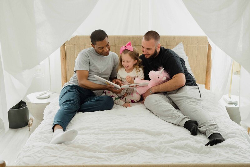 Two dads snuggle with their child, each holding either end of a children's book that they are reading together. Everyone is smiling happily.