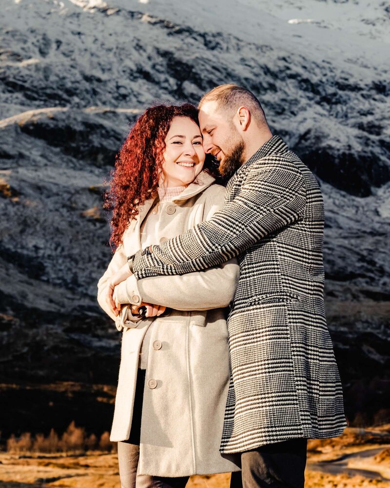 A couple warmly embracing in front of a dramatic mountain landscape, dressed in cozy neutral tones and smiling affectionately, captured by a UK wedding photographer.

