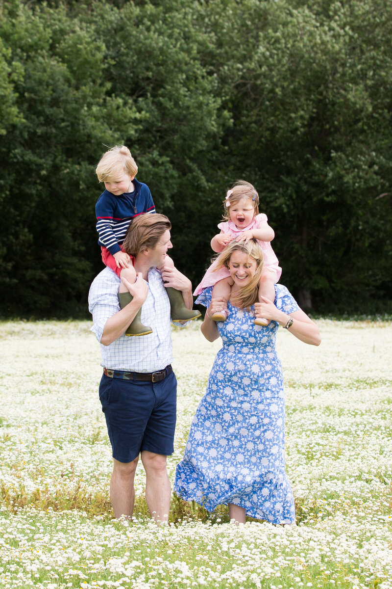 A vibrant family portrait in a field of wildflowers, with parents joyfully carrying their children on their shoulders, by Suzy Slemen Photography.
