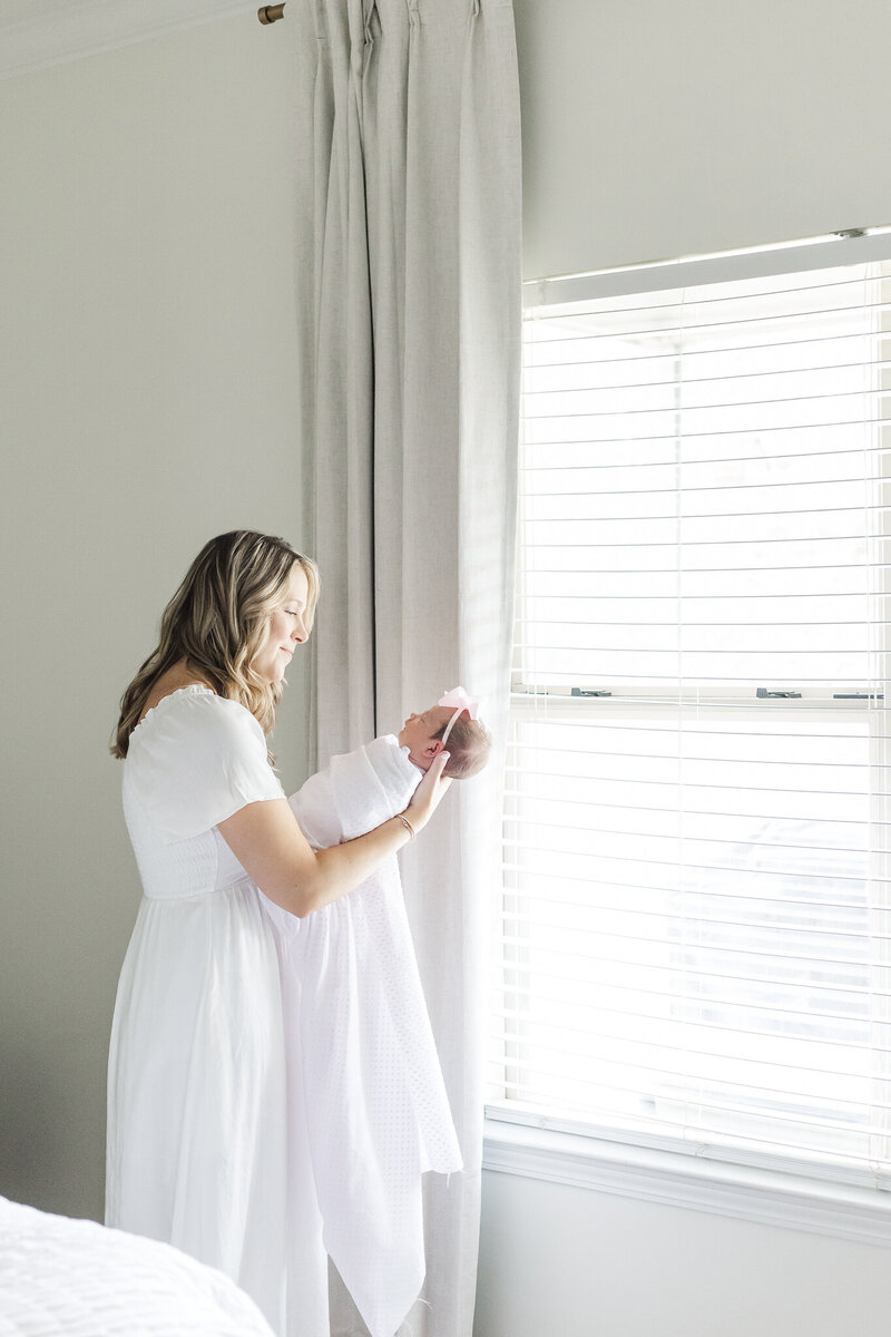 mom standing by window in a white dress smiling down at newborn baby girl wrapped in white swaddle during in-home newborn session with 5U Photography at their home in Alabaster, Alabama