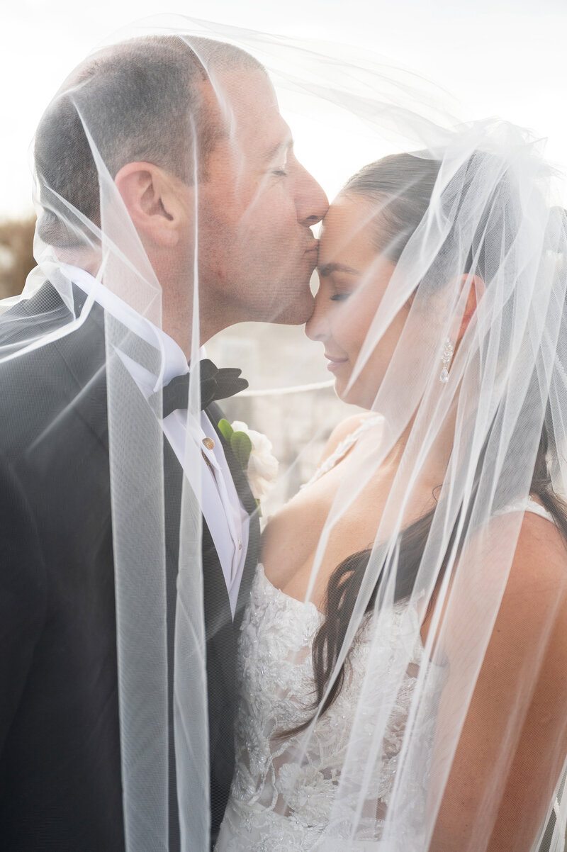 Groom kissing his bride on the forehead while under a veil