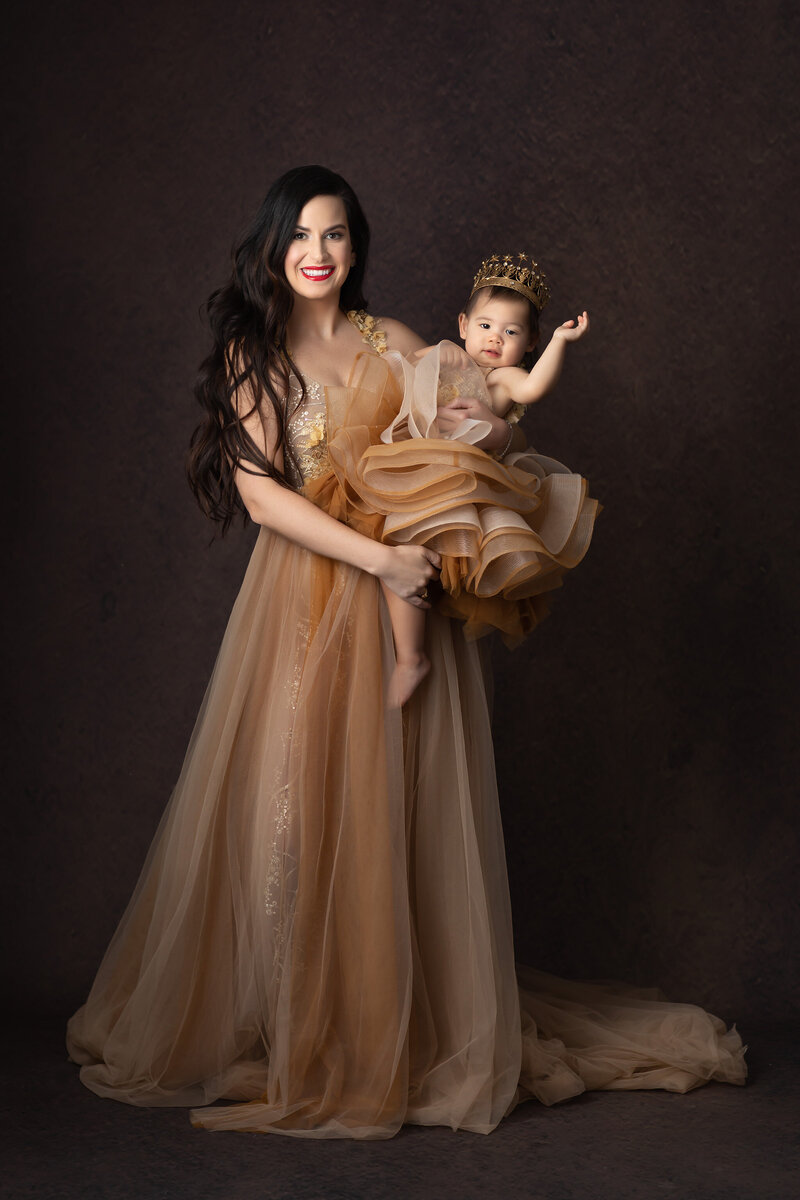A mother in a gold gown holds her baby girl wearing a matching dress and gold crown, both smiling in an elegant studio portrait. Captured by a Dallas Baby Milestone Photographer.