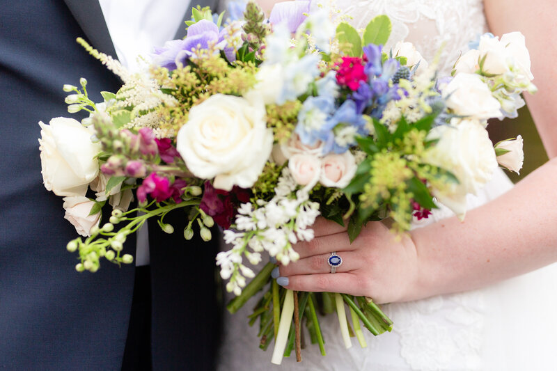 bride wears a sapphire engagement ring while holding blue and purple  bouquet