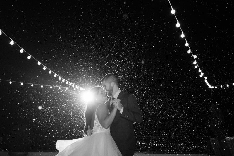 Black and white photograph of a bride and groom dancing intimately under twinkling string lights during a snowy evening at their wedding reception at the White Room in historic downtown St. Augustine. The couple is silhouetted against falling snow with dramatic backlighting creating a romantic, dreamy atmosphere.