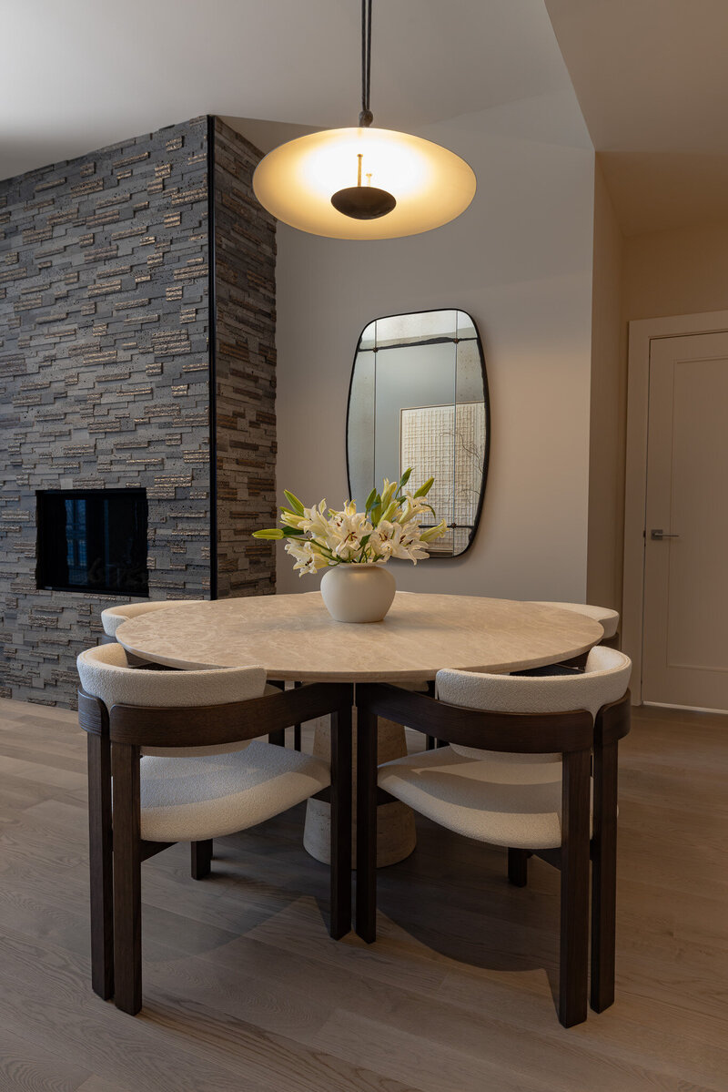 Vertical shot of a luxury dining room with a travertine table, white boucle chairs, and a stone fireplace in a Grand Hyatt Deer Valley Airbnb, designed by Sister Studio.
