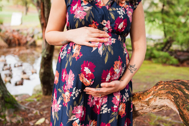 Maternity bump photo at Point Defiance Park in Tacoma WA focused on mom's hands on top and bottom of her growing belly in a flowered dark blue dress