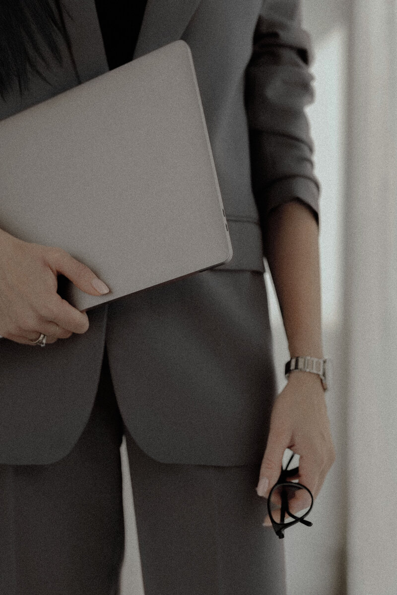 Close-up of a woman in a tailored gray suit holding a laptop and glasses, embodying confidence, sophistication, and modern business elegance.