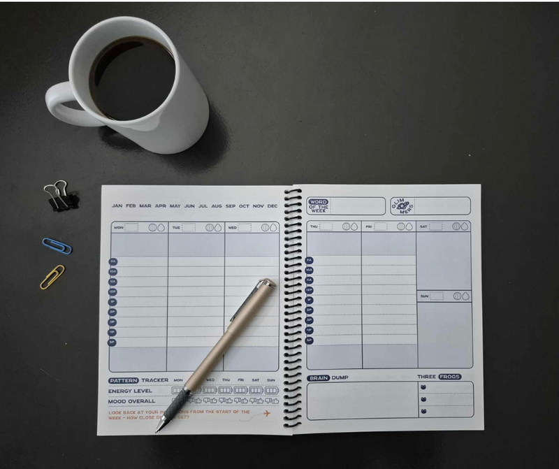 A flat lay of the Time Magic Planner open to a weekly spread on a dark table, with a cup of coffee, paper clips, and a pen placed across the pages.