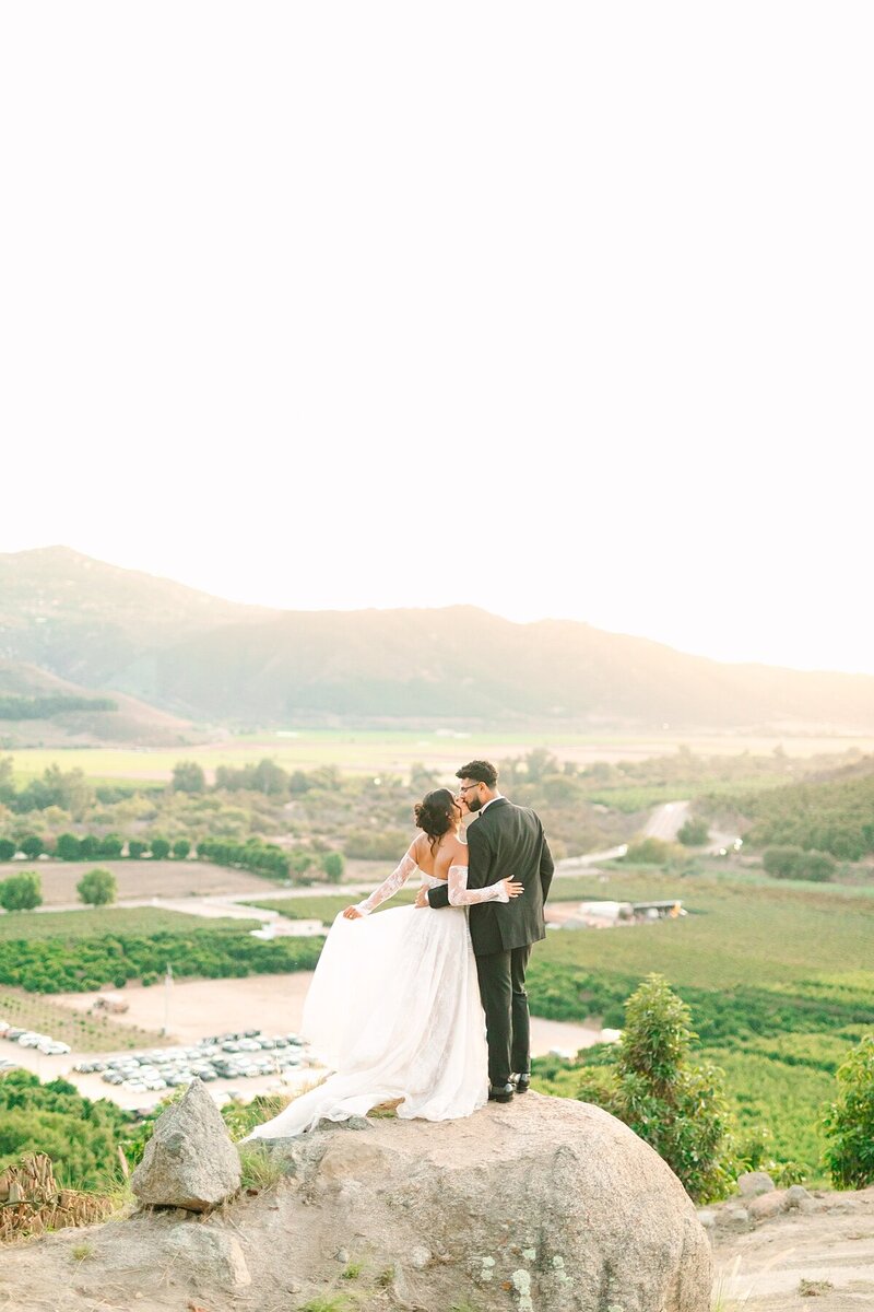 Bride and groom holding on to one another and kissing at the Eagle's Nest lookout point at Rancho Guijito Vineyard during their wedding pictures.