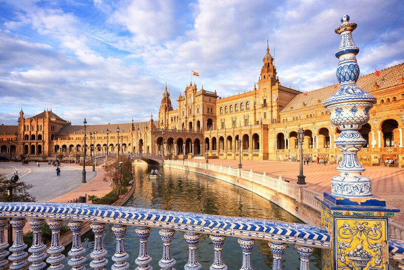 Plaza de España in Seville, Spain, featuring the grand semicircular building with towers, a canal, and a colorful ceramic balustrade in the foreground under a partly cloudy sky.