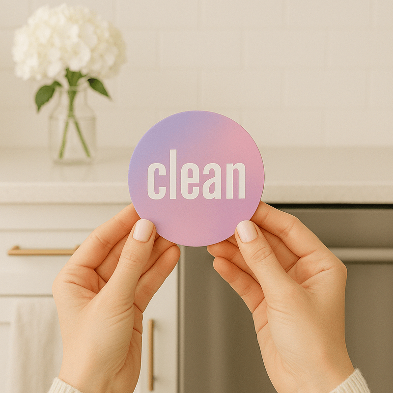 Two hands hold a pink and purple, round magnet with text that says "clean" in lowercase, bold, white letters with a elegant, minimalistic kitchen in the background