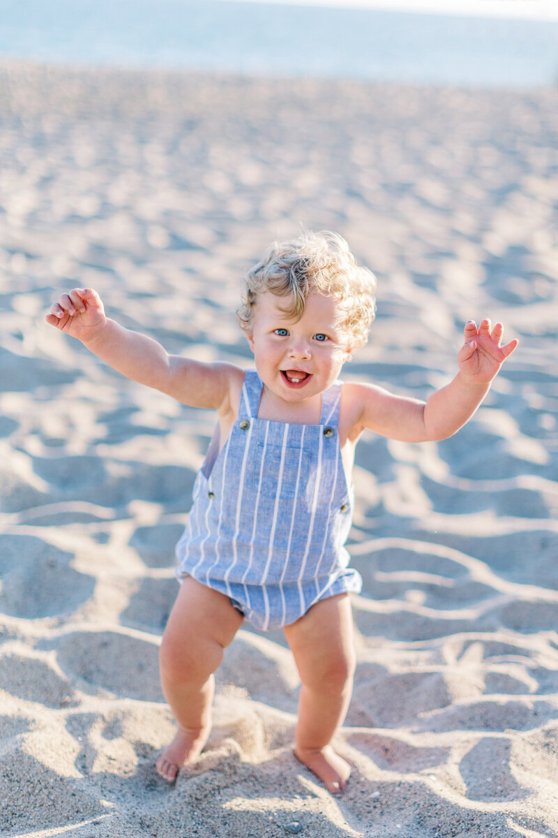 A baby boy in a blue jumper smiling on the beach in Camarillo