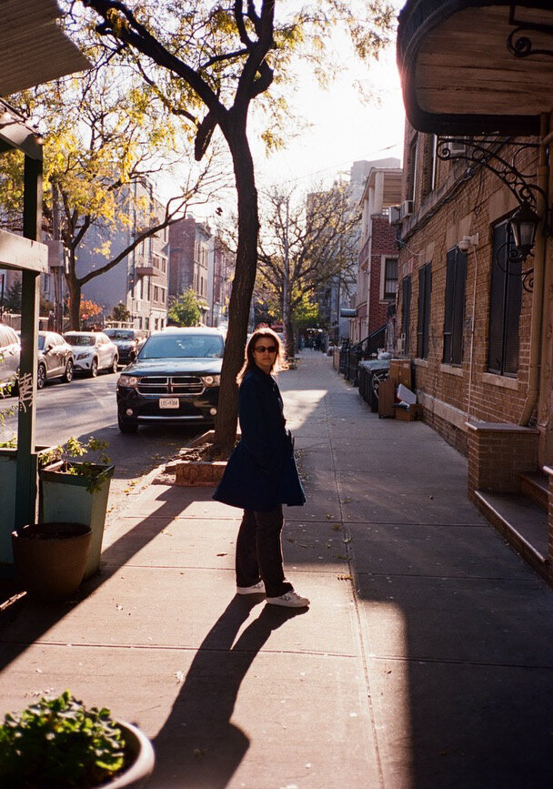 A woman wearing a navy coat and sunglasses walks along a sunlit New York City sidewalk lined with brownstone buildings, trees, and parked cars, capturing a peaceful urban moment in the afternoon light.