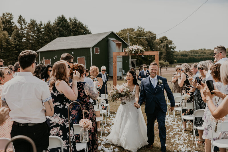 Bride and groom walk hand-in-hand through a field on their wedding day. Bride is smiling at the camera and the groom is looking over and smiling at the bride.