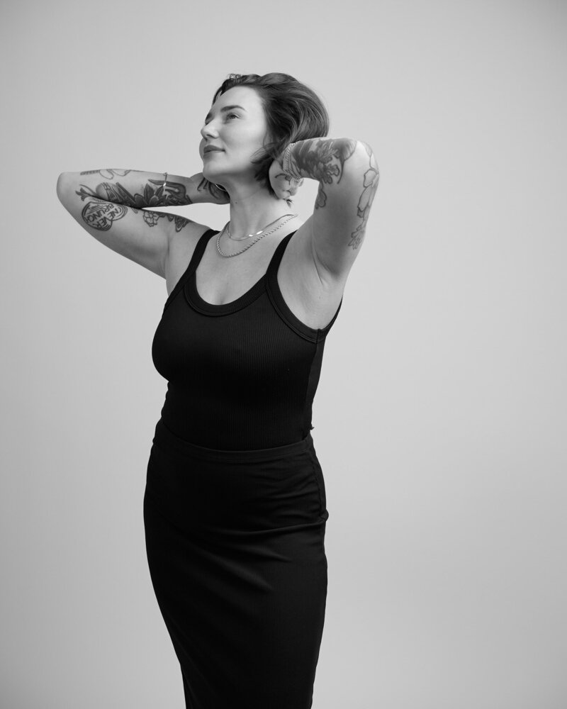 Black and white portrait of a tattooed woman standing with her hands resting behind her neck, gazing upward with a peaceful, confident expression. She wears a fitted black tank dress and layered necklaces, exuding strength and self-assurance. A striking, unfiltered moment from 'THIS IS ME – A Photography Experience by Katie Campbell' in Atlanta, GA, celebrating individuality and empowerment.