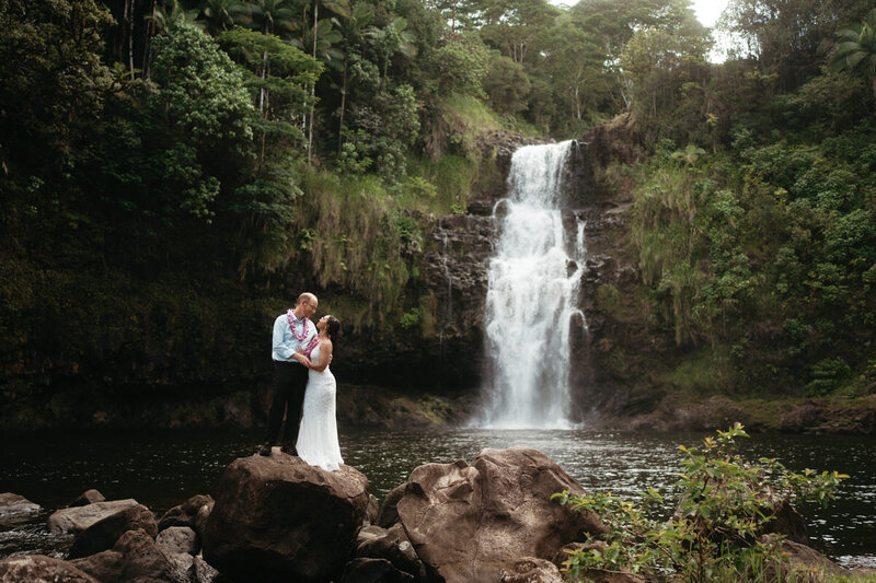 Eloping couple stands on top of a huge rock with a big waterfall behind them for an awesome Elopement Photo Idea