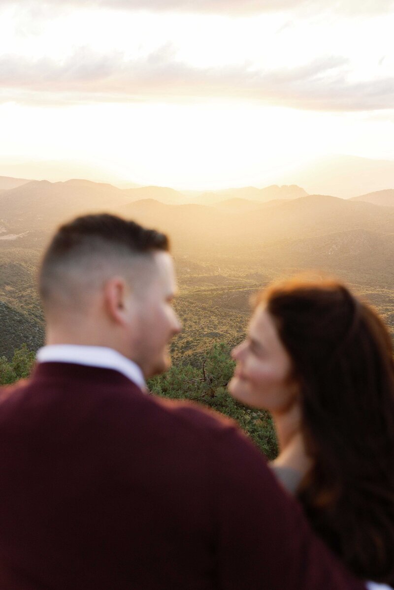 Couple looks at each other with a colorful sunset in the mountains behind them.
