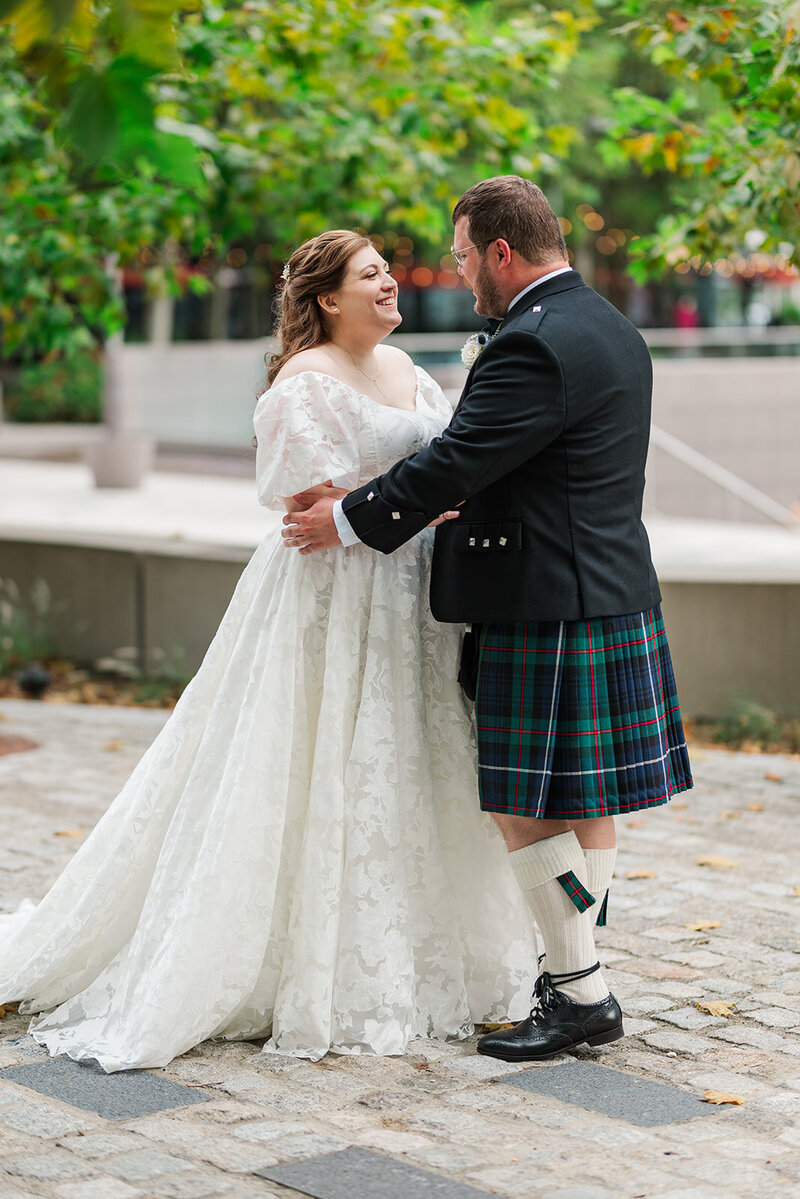 Jewish bride smiling and holding Scottish groom outside on a white brick paved walkway in Navy Yard DC