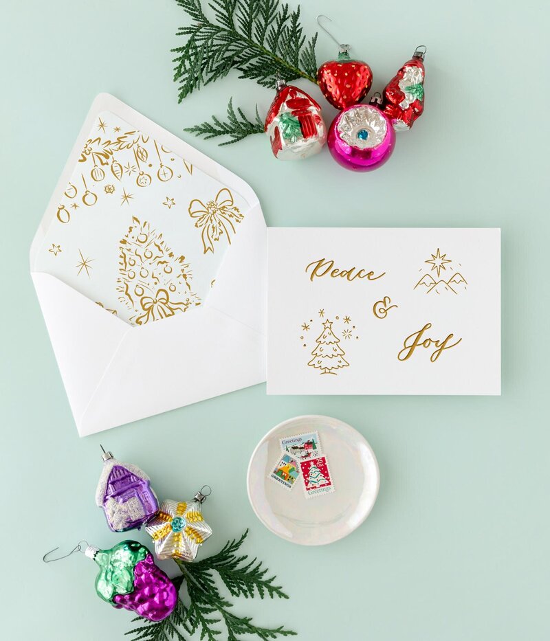 A greeting card in gold letterpress that says 'Peace & Joy' with matching envelope on a mint background with Christmas ornaments