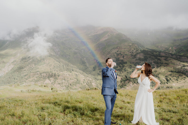 Bride and groom drink beers in front of a rainbow on a mountain top at their Ouray, Colorado Elopement