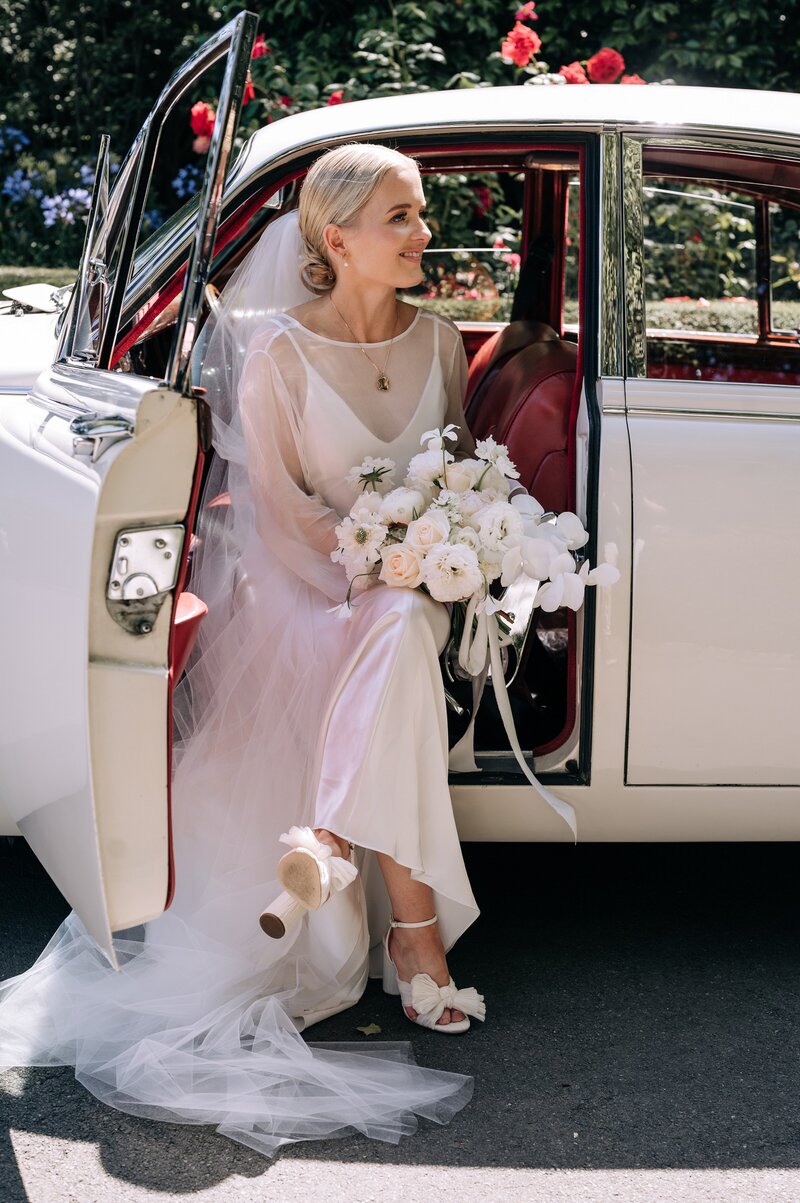 a bride in kenny harlow dress sits in a vintage white car with a white bouquet and ivory loeffler randall heels in christchurch