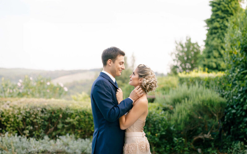 Couple is posing face to face holding each other surrounded by lush greenery.
