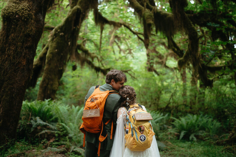 groom kisses bride on the head while they're wearing hiking backpacks in the forest | Elopement Photo Ideas