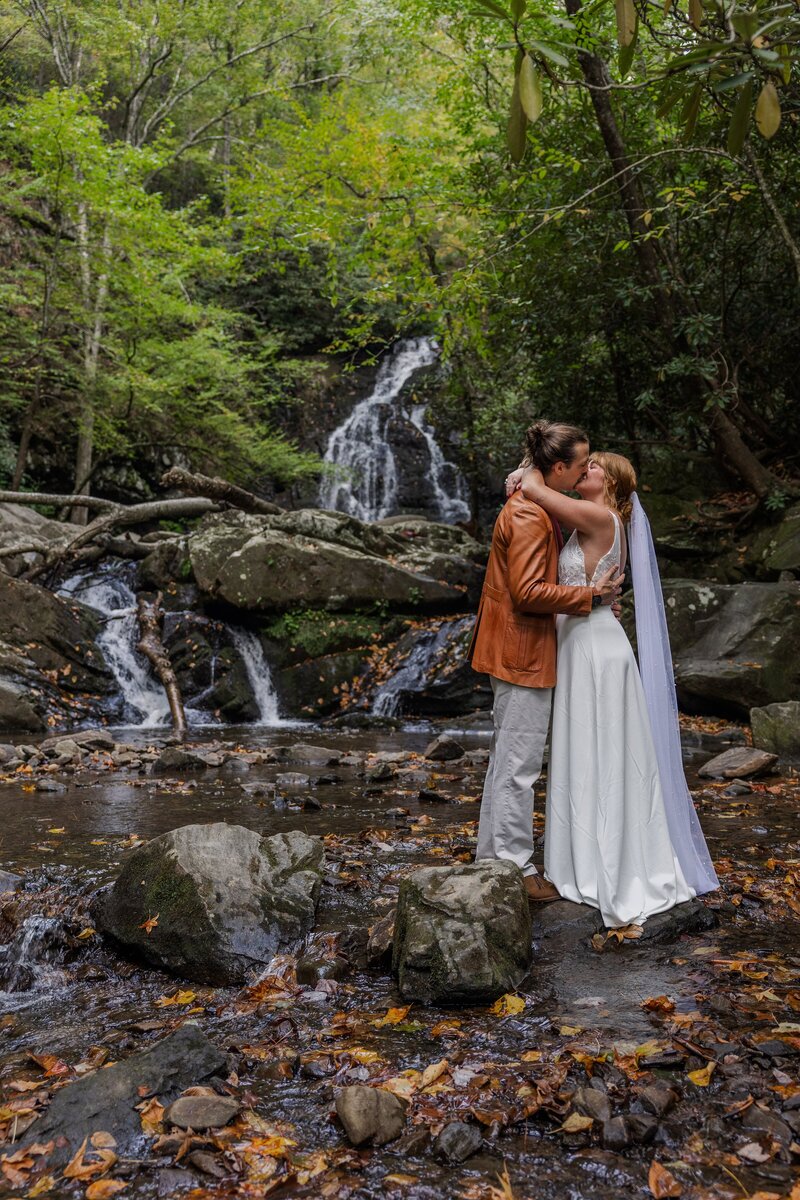 A bride and groom stand in front of a large, tumbling waterfall sharing a kiss. The bride is in a flowy wedding dress with a long veil and the groom is wearing tan pants with an orange leather jacket. They are surrounded by rocks and lush green trees.